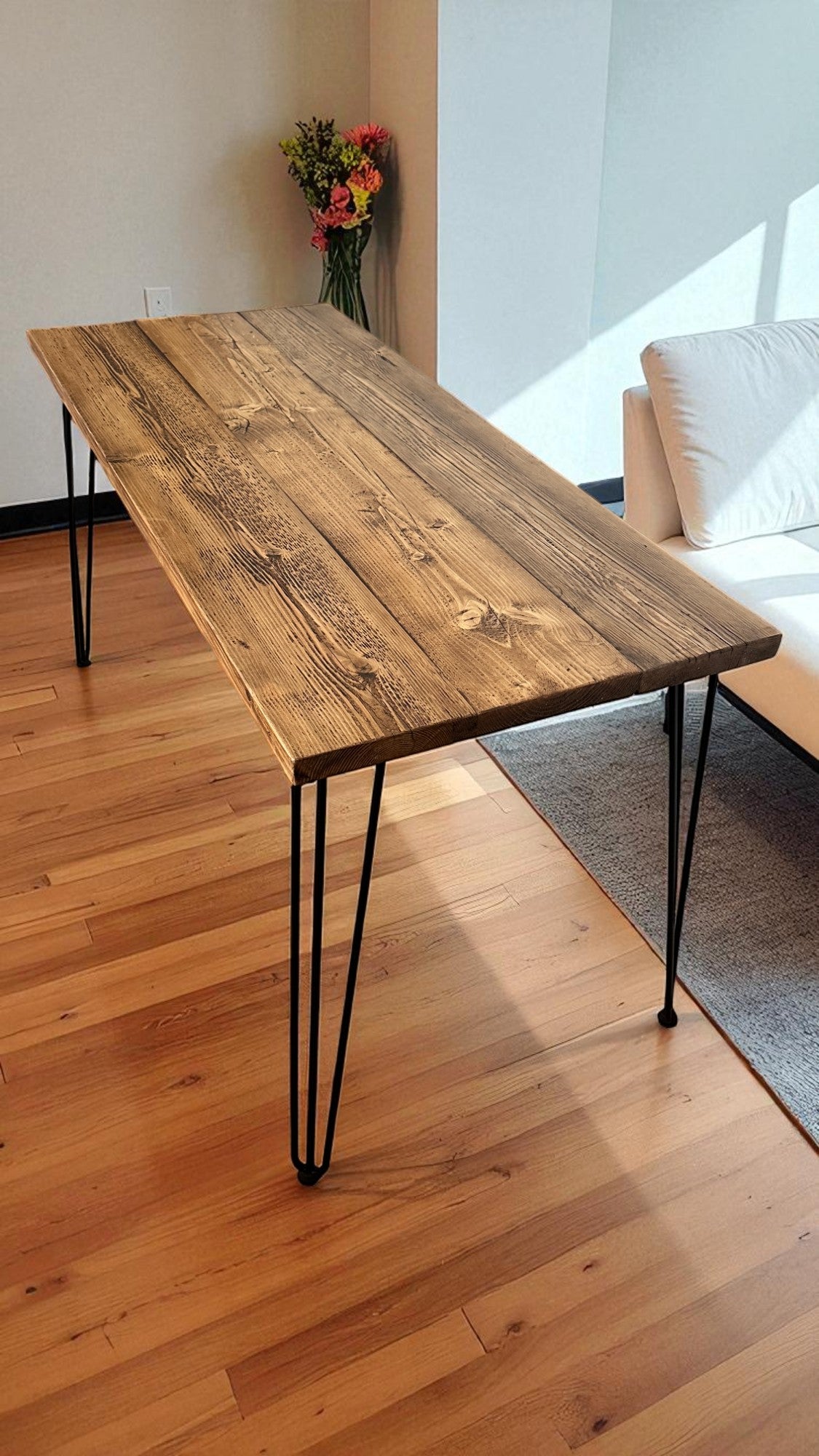 Wooden table with black hairpin legs in a room with a couch and flowers.
